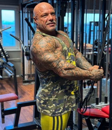 Ein muskulöser Mann mit tätowierten Armen trainiert in einem Fitnessstudio.